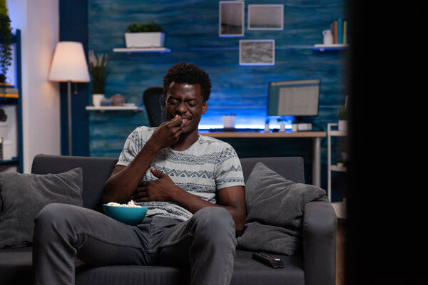 African american guy sitting on couch holding bowl with popcorn