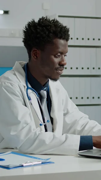 Médico afro-americano usando laptop enquanto sentado na mesa — Fotografia de Stock