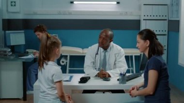 Afrikalı Amerikalı doktor küçük hasta ve annesiyle konuşuyor.