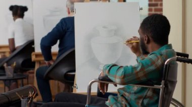 Tekerlekli sandalyedeki sanat sınıfı öğrencisi vazo çizmek için tuval üzerine kalem kullanıyor.