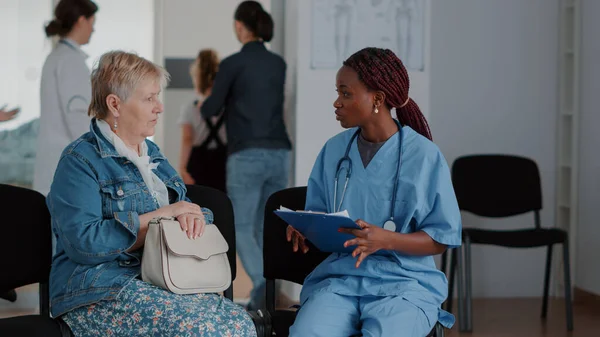 Afrikansk amerikansk sjuksköterska förklarar diagnos för äldre kvinna i väntrummet — Stockfoto