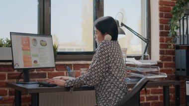 Bilgisayarda çalışırken ofis telefonunu kullanan Asyalı kişi