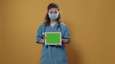 Tablet bilgisayarlı, yeşil ekranlı, hastane üniforması giyen ve stüdyoda koruyucu maske takan kadın portresi. Reklam alanı için dijital dokunmatik ekran aygıtı kullanan sağlıkçı.