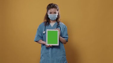 Kendine güvenen kafkas doktoru, elinde yeşil ekranlı dijital tablet, hastane üniforması ve kovid koruma maskesi ile stüdyoda poz veriyor. Tıbbi konsepti tanıtmak için dokunmatik ekran aygıtı kullanan sıhhiye.