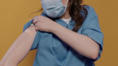 Stüdyoda cerrahi maske takan, kollarını kaldıran ve grip aşısından sonra yara bandı gösteren doktorların yakın çekimi. Üniformalı sıhhiyenin detaylı görüntüsü, bağışıklık noktasını kapatan bandajı gösteriyor..