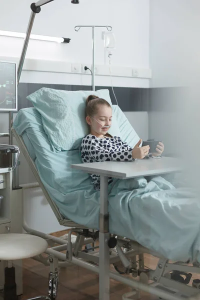 Menina doente brincando no smartphone enquanto descansa na cama do paciente na sala de clínica pediátrica. — Fotografia de Stock