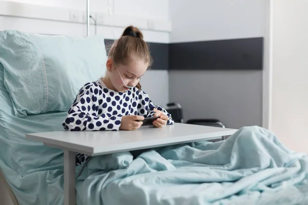 Хвора дівчинка грає на смартфоні, відпочиваючи на ліжку пацієнта в педіатричній клінічній кімнаті . — стокове фото