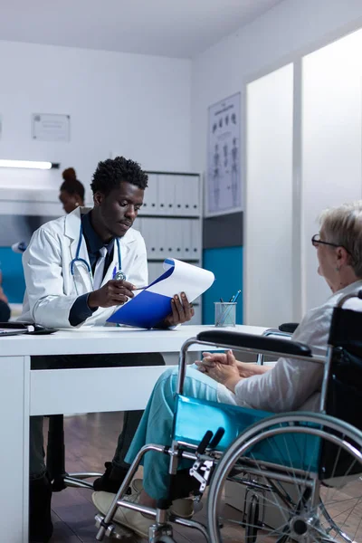 Klinikarzt überprüft Untersuchungsergebnisse von Seniorin im Rollstuhl. — Stockfoto