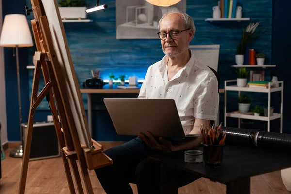 Artiste senior assis devant un chevalet tenant un ordinateur portable regardant la galerie d'art en ligne à la recherche d'inspiration — Photo