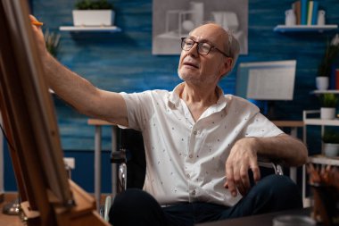 Tekerlekli sandalyede oturan yaşlı bir sanatçı tuvalde kalem kullanarak siyah beyaz çizim yapıyor.