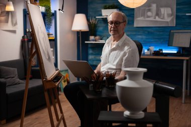 Üst düzey sanatçı elinde dizüstü bilgisayarla sanat stüdyosunda profesyonel çizim araçlarıyla oturuyor.