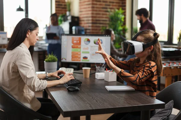 Playful little child wearing VR headset enjoys virtual reality technology while busy mother working at computer. — Stock Photo, Image