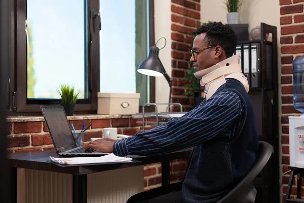 非洲裔美国人穿着颈项支架坐在创业办公室的办公桌前，在事故后康复 — 图库照片