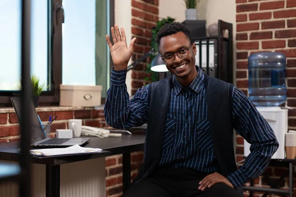 Dipendente startup sorridente con gli occhiali posa casuale agitando ciao seduto alla scrivania — Foto Stock