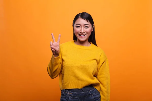 Šťastná krásná asijská žena dává mír znamení a zároveň se srdečně usmívá na oranžovém pozadí. — Stock fotografie