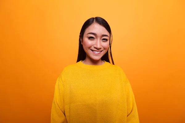 Belle femme asiatique heureuse souriant chaleureusement tout en portant un pull jaune sur fond orange. — Photo