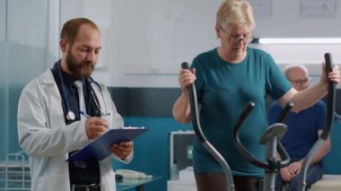 Doktor durağan bisikletteki yaşlı kadına tavsiye veriyor.