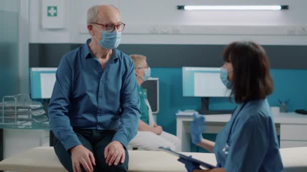 Stary pacjent z maską na twarzy rozmawiający z pielęgniarką — Wideo stockowe