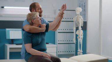 Erkek osteopat, yaşlı kadına kol kaslarını esnetmesi için yardım ediyor.