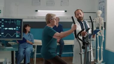 Erkek doktor yaşlı kadının sabit bisiklet kullanmasına yardım ediyor.