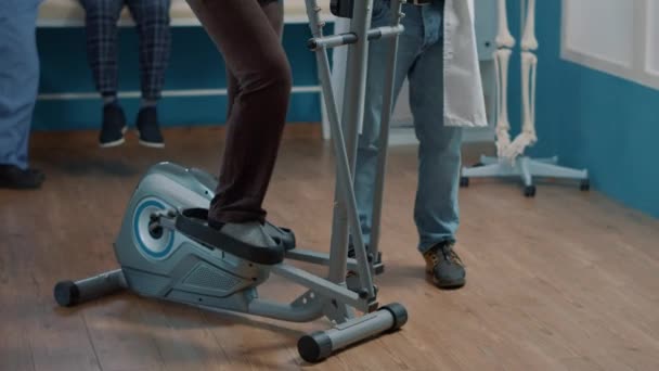 Old patient using stationary bicycle at physical therapy with physician — Stock Video