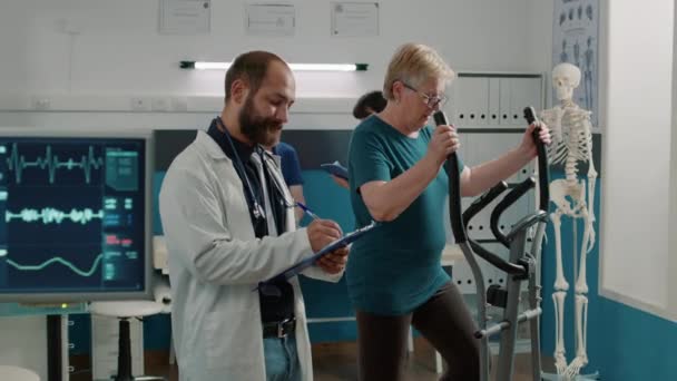 Osteopath macht Notizen und beobachtet Patienten bei der Nutzung von stationärem Fahrrad — Stockvideo