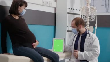 Yüzü maskeli hamile hasta ve doktor yeşil ekranlı tablet kullanıyor