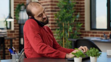 Genç yetişkin telefona cevap veriyor ve bilgisayarla uğraşıyor.