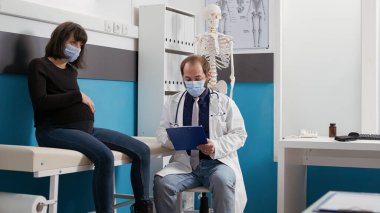 Yüzü maskeli hamile kadın ve doktor muayene ediyor.