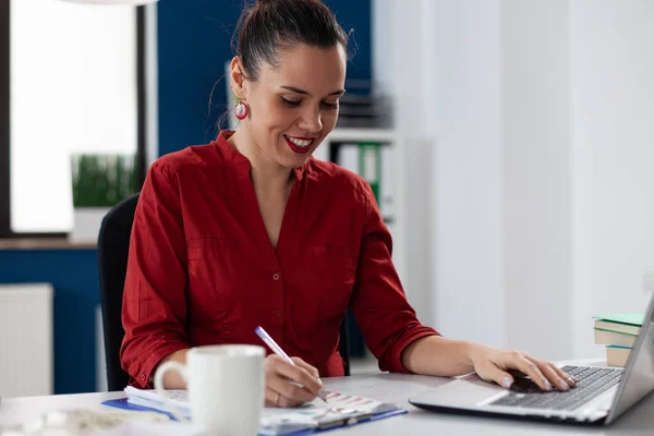 Businesswoman writing notes on clipboard with one hand on laptop.