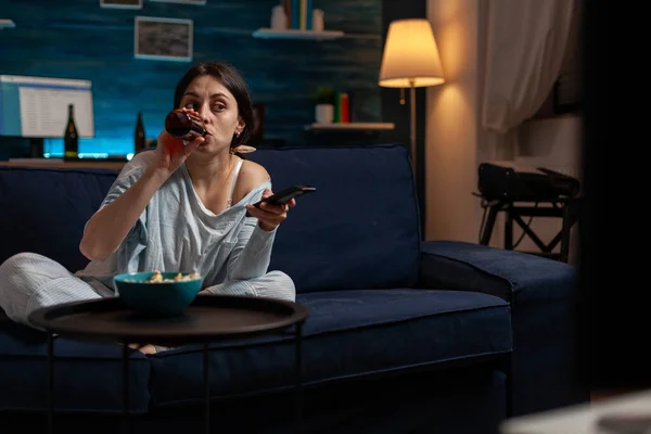 Молодая женщина пьет пиво из бутылки и переключает телеканалы — стоковое фото