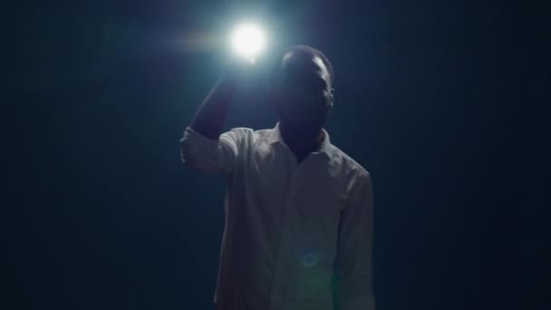 Jongeman zit in het donker en gebruikt zaklamp om rond te zoeken — Stockvideo