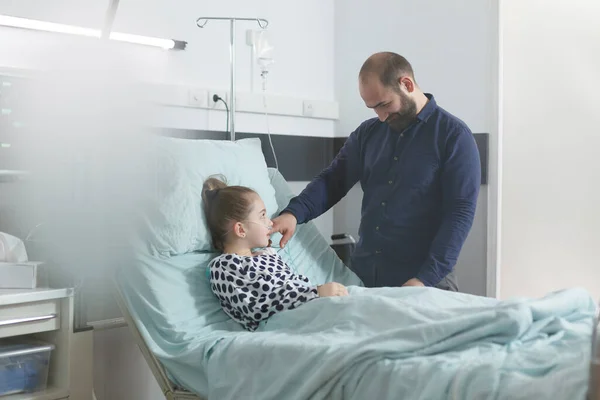 Padre atento visitando a la pequeña hija enferma en la clínica pediátrica mientras ofrece apoyo emocional. —  Fotos de Stock