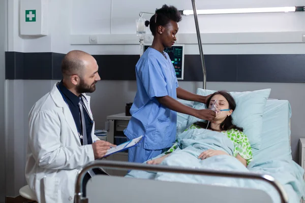 医生拿着剪贴板坐在床边时，护士给不省人事的女病人戴上氧气面罩 — 图库照片