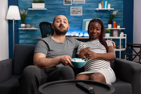 Cheerful interracial pregnant couple sharing a bowl of snacks