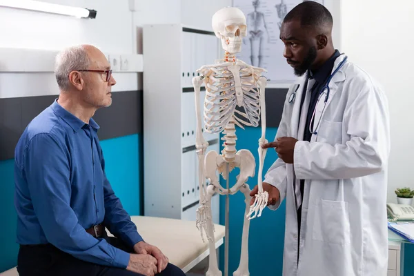 Médico fisioterapeuta mostrando esqueleto humano anatómico explicando dolor de espalda al paciente anciano —  Fotos de Stock