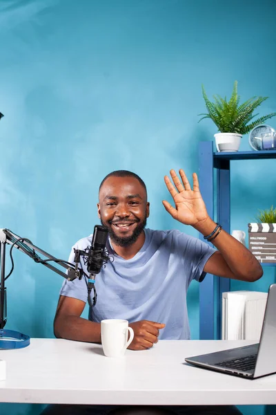 Portret afrykańskiego vloggera machającego ręką przed kamerą w studiu nagraniowym z profesjonalnym mikrofonem — Zdjęcie stockowe