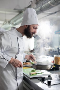 Yetenekli baş aşçı tavada spatulayla karıştırırken akşam yemeği için taze otlarla süslenmiş gurme yemeği pişiriyor..