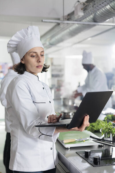 Restaurant kitchen worker with laptop searching gourmet cuisine dinner dish for cooking contest held at restaurant.