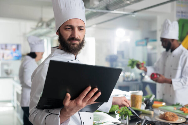 Food industry worker having laptop, following gourmet dish recipe on screen stirring ingredients in pan. Head chef with computer preparing garnish for main course while in restaurant kitchen.