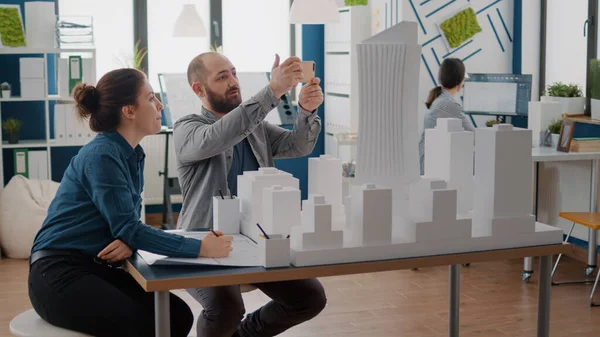 Arbetskamrater som använder mobiltelefon för att utforma konstruktionslayout efter byggnadsmodell på bordet. — Stockfoto