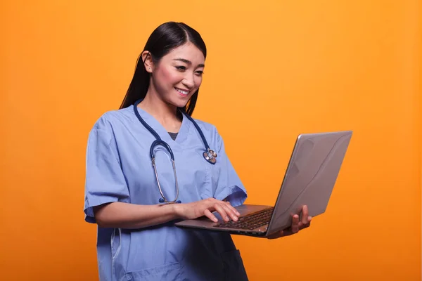 Счастливая улыбающаяся медсестра азиатской клиники, носящая стетоскоп и пользующаяся современным ноутбуком. — стоковое фото