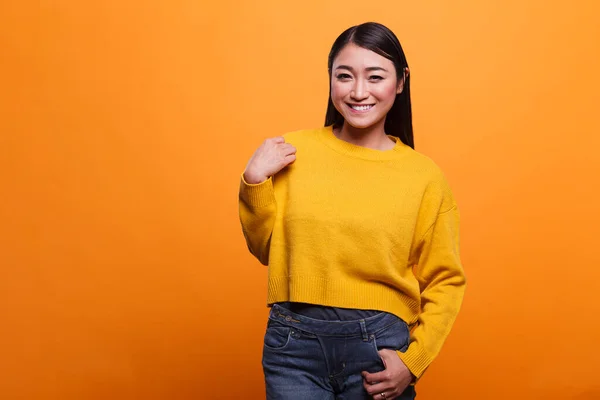 Portrait of positive friendly smiling heartily asian woman wearing yellow sweater on orange background.