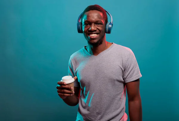 Young guy holding cup of coffee and enjoying music on headset — Stock Photo, Image