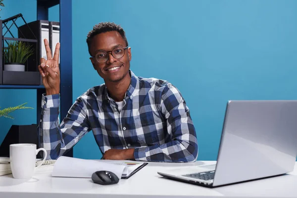 Sonriente hombre alegre haciendo símbolo ganador con la mano mientras está en el espacio de trabajo de la oficina. —  Fotos de Stock