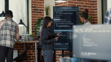 Software engineer holding digital tablet analyzing code on wall screen tv explaining errors to colleague programer