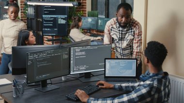 Programcı çalışma masasında birden fazla ekranla oturuyor. Çalışma arkadaşıyla konuşuyor.