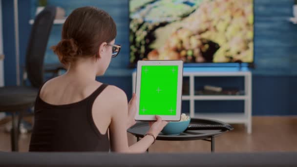 Mujer joven sentada en un sofá sosteniendo una tableta digital vertical con pantalla verde viendo contenido de redes sociales en línea — Vídeos de Stock