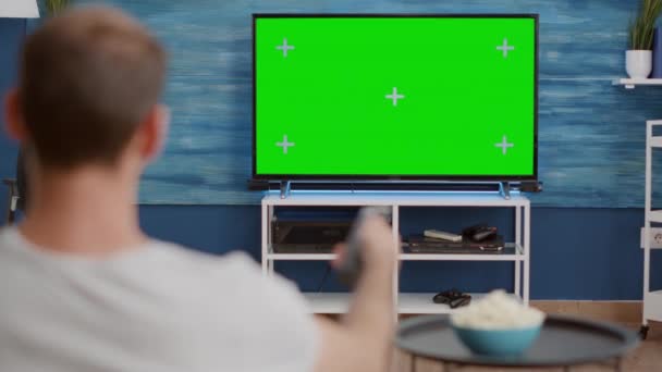 Над видом на плече людини, що перемикає канали, дивлячись на зелений екран по телевізору і сидячи на дивані — стокове відео