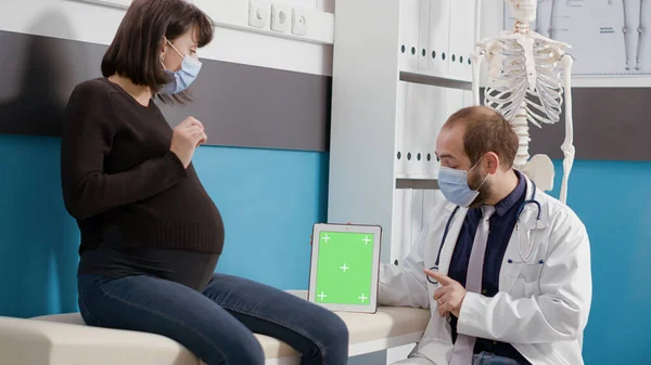 Yüzü maskeli hamile hasta ve doktor yeşil ekranlı tablet kullanıyor — Stok fotoğraf